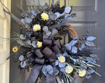 May include: A decorative wreath with a dark color palette, featuring black and gray leaves, berries, and accents. The wreath includes a large black bow and small yellow flowers. It is displayed on a black door.