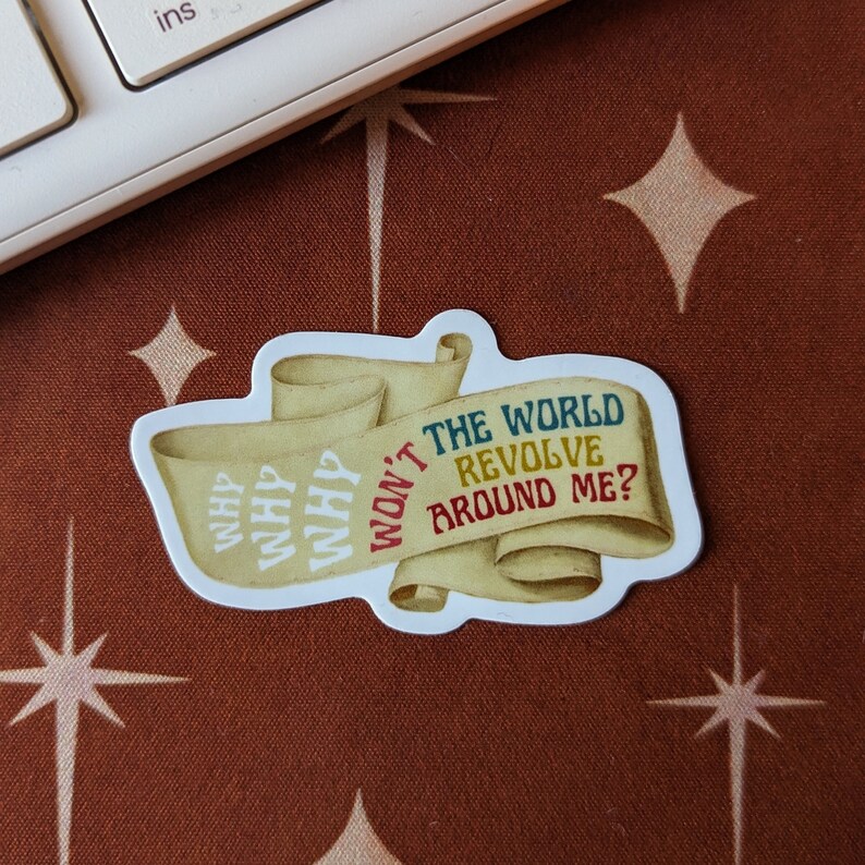May include: A sticker with a scroll design. The scroll reads "THE WORLD REVOLVE AROUND ME?" in colorful text. The left side of the scroll has the word "WHY" repeated three times. The sticker is on a brown surface.