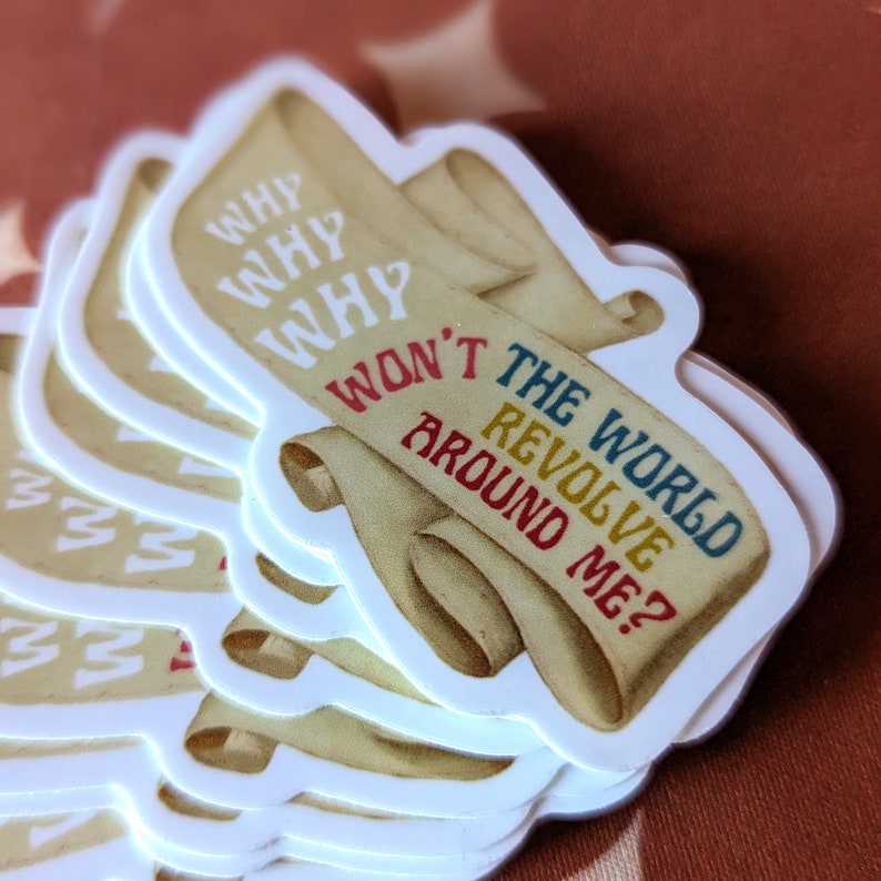 May include: A stack of stickers with a scroll design. The scroll has the text "WHY WHY WHY WON'T THE WORLD REVOLVE AROUND ME?" in various colors. The stickers have a white border and a tan background.