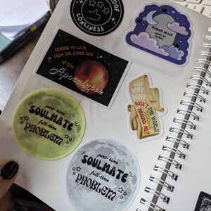 May include: A collection of colorful stickers on a spiral notebook. The stickers feature various designs, including a black circle with the text "Bulletproof Lonliness", a moon with the words "Soulmate Problem", and a tombstone with the text "Buried Alive Inside My Dreams".