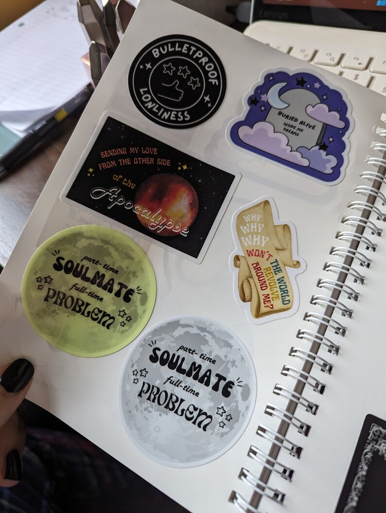 May include: A collection of colorful stickers on a spiral notebook. The stickers feature various designs, including a black circle with the text "Bulletproof Lonliness", a moon with the words "Soulmate Problem", and a tombstone with the text "Buried Alive Inside My Dreams".