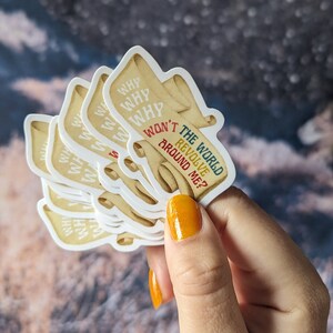 May include: A stack of stickers with a scroll design and the text "WHY WON'T THE WORLD REVOLVE AROUND ME?" in various colors. The stickers have a cream-colored background and are held in a hand with orange nail polish.