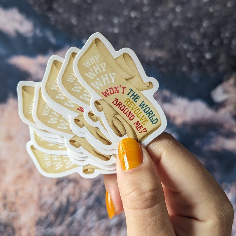 May include: A stack of stickers with a scroll design and the text "WHY WON'T THE WORLD REVOLVE AROUND ME?" in various colors. The stickers have a cream-colored background and are held in a hand with orange nail polish.