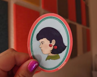 May include: An oval sticker featuring a profile illustration of a person with dark hair and cherry-shaped earrings. The sticker has a light blue background, a decorative teal scalloped border, and a coral-colored outer edge with small white text.