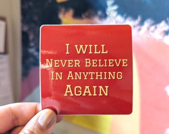 Könnte beinhalten: Roter quadratischer Aufkleber mit dem Text "I WILL NEVER BELIEVE IN ANYTHING AGAIN" in goldenen Buchstaben.