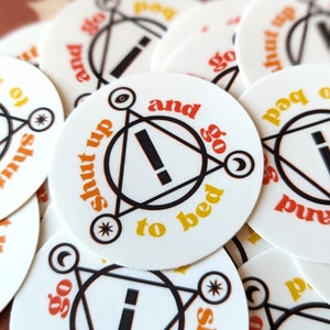 May include: A white sticker with a black triangle and a black exclamation point inside. The text "Shut up and go to bed" is written in a yellow and orange font around the triangle.