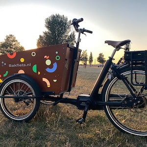 Op de afbeelding: Een zwarte elektrische bakfiets met een bruine houten bak. De bak heeft een kleurrijk geometrisch patroon en de tekst "Bakfiets.nl" erop gedrukt. De fiets heeft een zwart leren zadel en een zwarte accu.