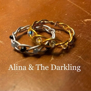 Peut inclure: Deux anneaux entrelacés, l'un en argent et l'autre en or, avec des perles noires et blanches et une pierre bleue. Les anneaux sont sur une surface brune. Alina & The Darkling.