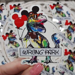 May include: A colorful watercolor sticker of a Tyrannosaurus Rex wearing Mickey Mouse ears and sunglasses, holding a red balloon. The dinosaur is standing on a sign that says "WRONG PARK".