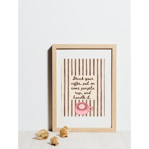 May include: A framed print featuring a beige background with vertical brown stripes. The text reads, "Drink your coffee, put on some gangsta rap, and handle it." Below the text is a small illustration of a pink coffee mug with a smiley face.