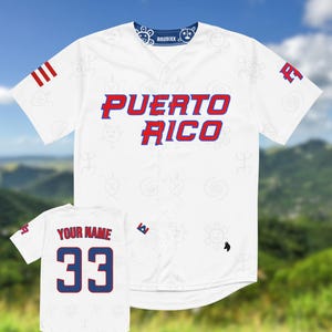 Puede incluir: Camiseta de b&eacute;isbol blanca con "PUERTO RICO" en letras rojas y azules. La camiseta tiene un cuello azul con dise&ntilde;os blancos y la marca "ANUBIXX". Una camiseta m&aacute;s peque&ntilde;a muestra "YOUR NAME" y el n&uacute;mero "33" en rojo y azul.