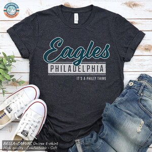 May include: A dark charcoal heather gray short-sleeve t-shirt featuring the word Eagles in a teal script font, with the word PHILADELPHIA in white block letters below, and the text IT'S A PHILLY THING printed in smaller white letters.