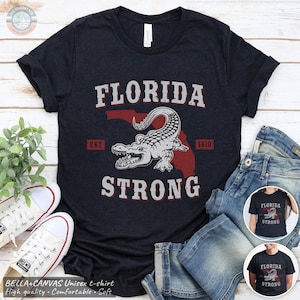 May include: A dark blue t-shirt with a white alligator graphic and the text "Florida Strong" in white. The alligator is superimposed on a red outline of the state of Florida. The text "Est 1819" is in white below the alligator.