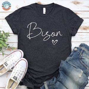 May include: A dark charcoal gray short-sleeve t-shirt featuring the word Bison in a white cursive script font with a small white heart outline underneath. The shirt is displayed alongside a pair of blue denim jeans and white sneakers.