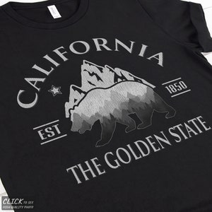 May include: A black t-shirt featuring a white graphic print of a bear silhouette filled with a mountain and forest landscape. The text reads CALIFORNIA, EST 1850, and THE GOLDEN STATE, accompanied by a small star icon.