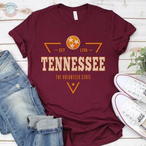 May include: A maroon short-sleeve t-shirt featuring a graphic with the text TENNESSEE, THE VOLUNTEER STATE, EST 1796, and a circular emblem containing three white stars. The design includes orange geometric lines and a small star.