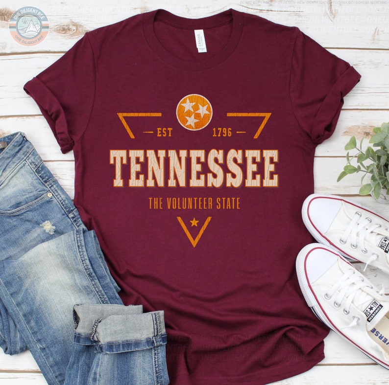 May include: A maroon short-sleeve t-shirt featuring a graphic with the text TENNESSEE, THE VOLUNTEER STATE, EST 1796, and a circular emblem containing three white stars. The design includes orange geometric lines and a small star.