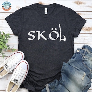 May include: A dark charcoal gray short-sleeve t-shirt featuring the white distressed text SKÖL printed across the chest, with the letter L stylized as a Viking axe. The shirt is paired with blue denim jeans and white canvas sneakers.