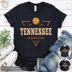 May include: A dark navy blue short-sleeve t-shirt featuring a distressed orange and white graphic. The design includes a circular emblem with three stars, the text TENNESSEE, EST 1796, and THE VOLUNTEER STATE, with a star below.