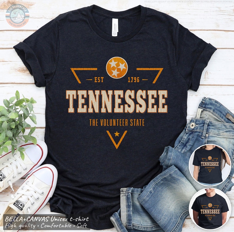 May include: A dark navy blue short-sleeve t-shirt featuring a distressed orange and white graphic. The design includes a circular emblem with three stars, the text TENNESSEE, EST 1796, and THE VOLUNTEER STATE, with a star below.