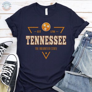 May include: A navy blue t-shirt featuring a distressed orange and white graphic that reads TENNESSEE THE VOLUNTEER STATE EST 1796. The design includes the Tennessee state flag emblem with three stars inside a circle and geometric accents.