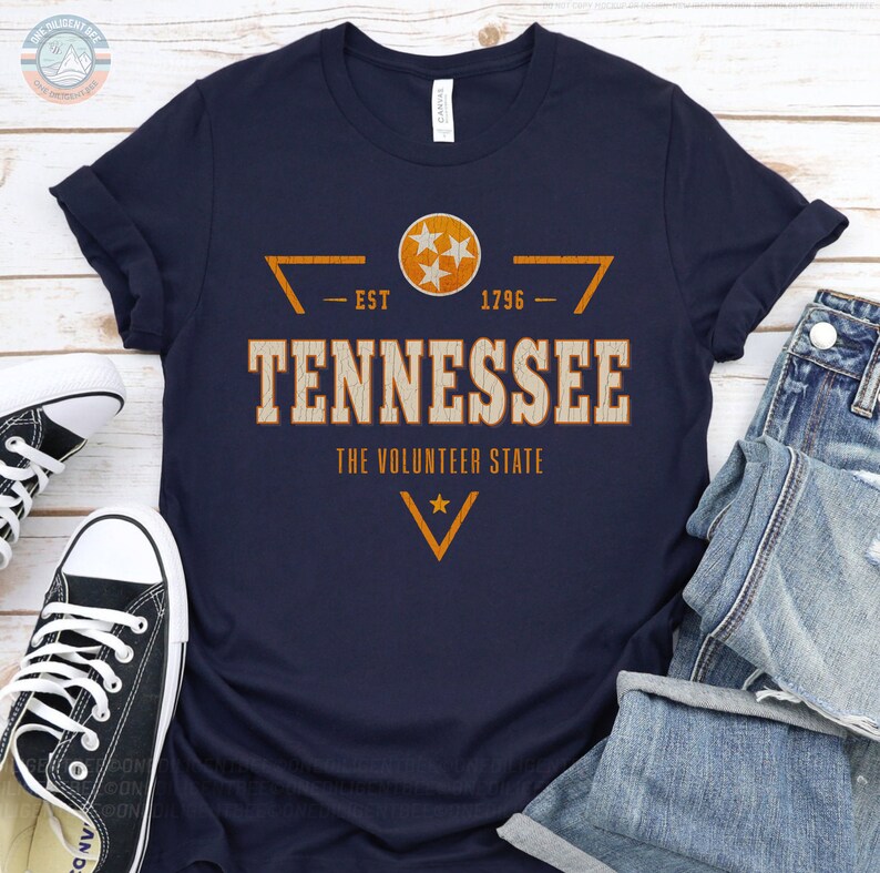May include: A navy blue t-shirt featuring a distressed orange and white graphic that reads TENNESSEE THE VOLUNTEER STATE EST 1796. The design includes the Tennessee state flag emblem with three stars inside a circle and geometric accents.