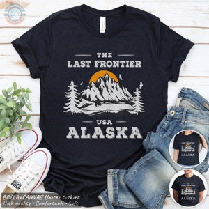 May include: A dark blue t-shirt with a white graphic design featuring a mountain range, a bear, and the text "The Last Frontier - USA - Alaska".
