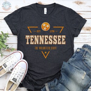 May include: A dark heather grey short-sleeve t-shirt featuring a distressed orange and white graphic that reads TENNESSEE THE VOLUNTEER STATE EST 1796 with a circular emblem containing three stars and a V-shaped chevron design.