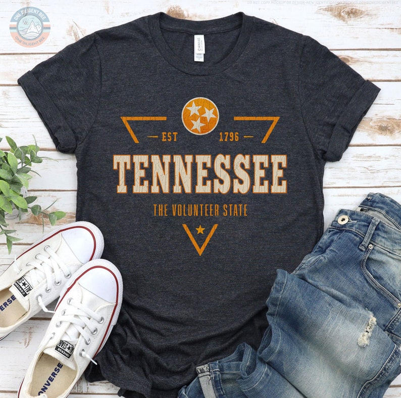 May include: A dark heather grey short-sleeve t-shirt featuring a distressed orange and white graphic that reads TENNESSEE THE VOLUNTEER STATE EST 1796 with a circular emblem containing three stars and a V-shaped chevron design.
