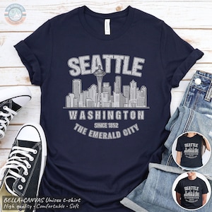 May include: A navy blue t-shirt featuring a white graphic of the Seattle skyline, including the Space Needle. The text reads SEATTLE, WASHINGTON, SINCE 1852, and THE EMERALD CITY. The design has a distressed, weathered appearance.