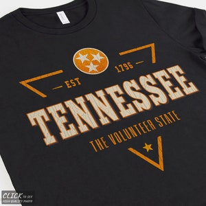 May include: A black t-shirt featuring a distressed orange and white graphic that reads TENNESSEE THE VOLUNTEER STATE, EST 1796, and includes a circular emblem with three stars, all printed in a weathered, cracked texture style.