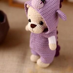 May include: A crocheted plush toy in the shape of a bear wearing a purple dinosaur costume. The bear has a brown nose and black eyes. The dinosaur costume has a hood with a purple spike on top and a tail.