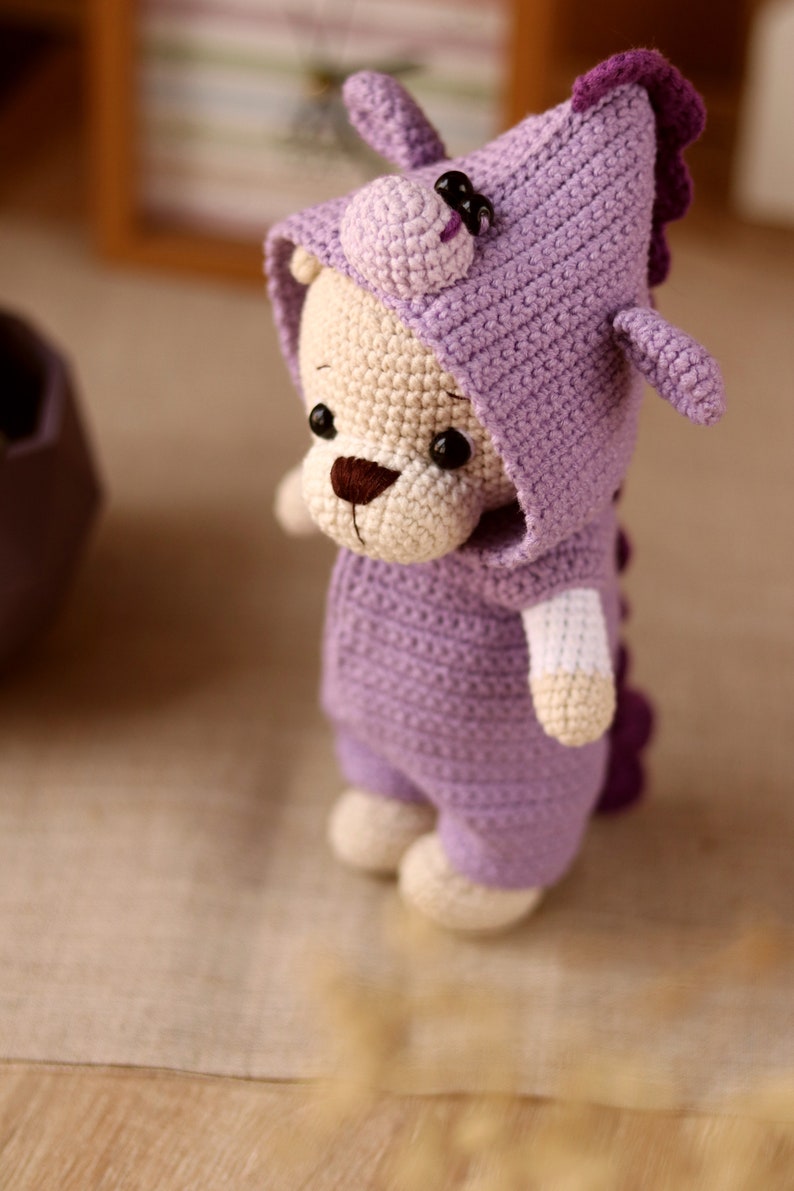 May include: A crocheted plush toy in the shape of a bear wearing a purple dinosaur costume. The bear has a brown nose and black eyes. The dinosaur costume has a hood with a purple spike on top and a tail.