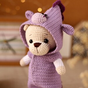 May include: A crocheted plush toy bear wearing a purple hooded outfit with a small unicorn horn on top of the hood.