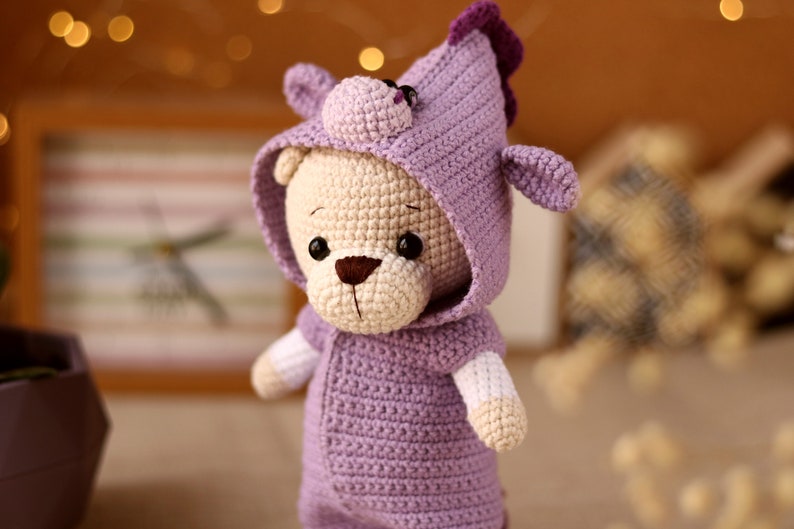 May include: A crocheted plush toy bear wearing a purple hooded outfit with a small unicorn horn on top of the hood.