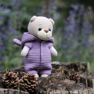 May include: A crocheted plush toy bear wearing a purple hooded jumpsuit. The bear is sitting on a tree stump with pine cones.