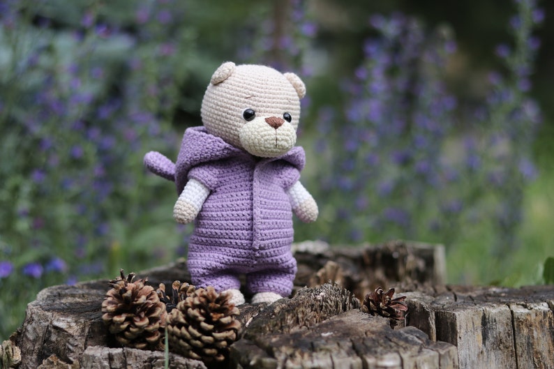 May include: A crocheted plush toy bear wearing a purple hooded jumpsuit. The bear is sitting on a tree stump with pine cones.