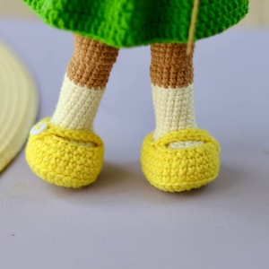 K&ouml;nnte beinhalten: Ein Paar gelbe geh&auml;kelte Schuhe mit wei&szlig;en Akzenten und einem kleinen wei&szlig;en Knopf vorne. Die Schuhe werden von einer geh&auml;kelten Puppe getragen.