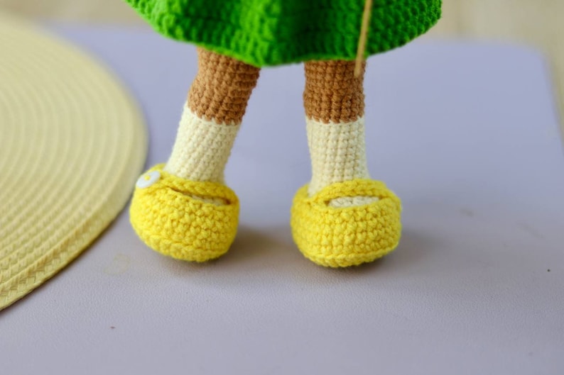 K&ouml;nnte beinhalten: Ein Paar gelbe geh&auml;kelte Schuhe mit wei&szlig;en Akzenten und einem kleinen wei&szlig;en Knopf vorne. Die Schuhe werden von einer geh&auml;kelten Puppe getragen.