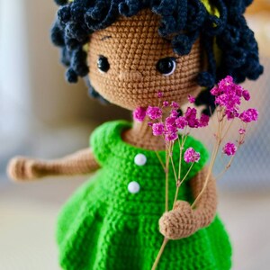K&ouml;nnte beinhalten: Eine geh&auml;kelte Puppe mit brauner Haut, schwarzen lockigen Haaren und einem gelben Stirnband. Die Puppe tr&auml;gt ein gr&uuml;nes Kleid mit wei&szlig;en Kn&ouml;pfen und h&auml;lt einen Strau&szlig; rosa Blumen.