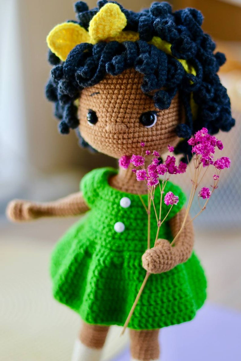 K&ouml;nnte beinhalten: Eine geh&auml;kelte Puppe mit brauner Haut, schwarzen lockigen Haaren und einem gelben Stirnband. Die Puppe tr&auml;gt ein gr&uuml;nes Kleid mit wei&szlig;en Kn&ouml;pfen und h&auml;lt einen Strau&szlig; rosa Blumen.