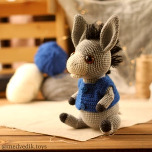May include: A gray crocheted donkey wearing a blue crocheted vest with two buttons. The donkey has brown eyes and a black mane.
