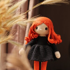 Peut inclure: Une poup&eacute;e en crochet avec des cheveux orange, une robe noire et des collants orange. La poup&eacute;e porte des chaussures violettes et se tient sur une surface brune.