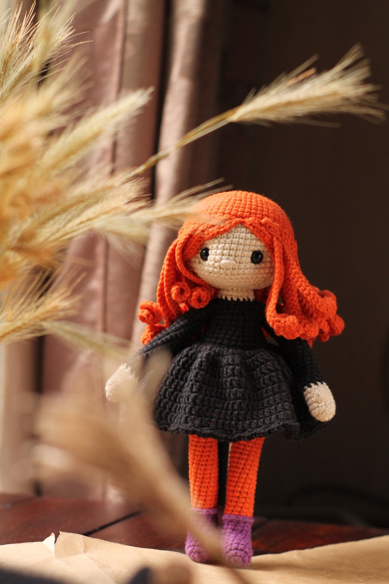 Peut inclure: Une poup&eacute;e en crochet avec des cheveux orange, une robe noire et des collants orange. La poup&eacute;e porte des chaussures violettes et se tient sur une surface brune.