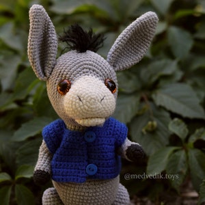 May include: A gray crocheted donkey wearing a blue crocheted vest with buttons. The donkey has brown eyes and a black mane.