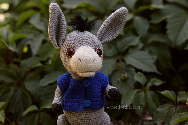 May include: A gray crocheted donkey wearing a blue crocheted vest with buttons. The donkey has brown eyes and a black mane.