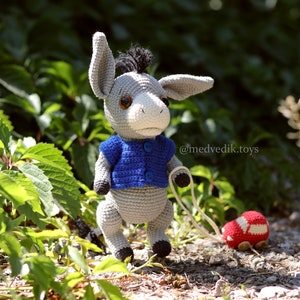 May include: A gray crocheted donkey toy wearing a blue crocheted sweater and pulling a red crocheted toy car. The donkey has brown eyes and a black mane. The toy is on a bed of green leaves and brown gravel. The text '@medvedik.toys' is visible in the image.