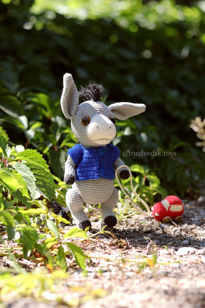 May include: A gray crocheted donkey toy wearing a blue crocheted sweater and pulling a red crocheted toy car. The donkey has brown eyes and a black mane. The toy is on a bed of green leaves and brown gravel. The text '@medvedik.toys' is visible in the image.