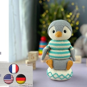 May include: A crocheted penguin toy with a grey body, white and turquoise striped torso, and orange beak. The penguin is sitting on a stack of crocheted rings, with a white and turquoise base.