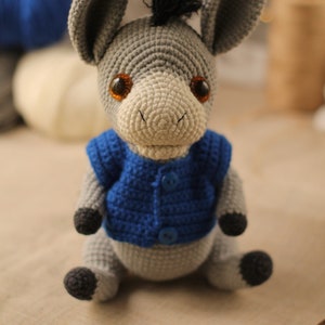 May include: A gray crocheted donkey wearing a blue crocheted vest with two buttons. The donkey has brown eyes and a black mane.